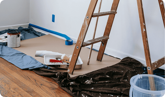 A room being prepped for painting with a ladder, paint cans, drop cloths, and blue painter's tape.