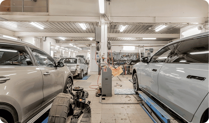 An auto repair shop with two cars in the foreground, one on a lift and another with a removed tire.