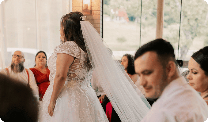 A bride in a white lace dress and veil stands with guests watching from the foreground and background.