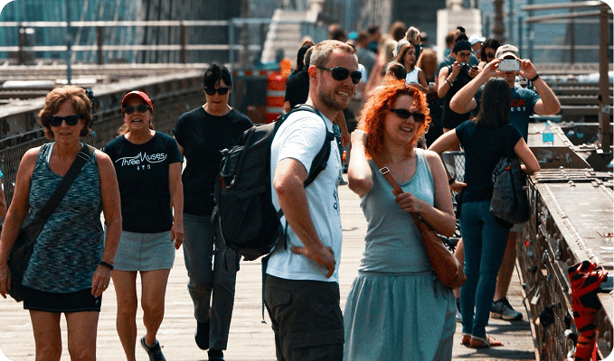 A diverse crowd of people, some smiling and wearing sunglasses, walk along a bridge on a sunny day.