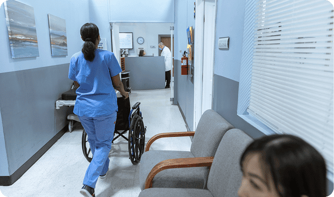 A nurse in blue scrubs pushes an empty wheelchair down a hospital hallway past waiting room chairs.