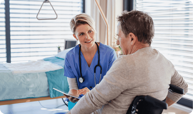 A smiling nurse in blue scrubs holds a tablet while talking to a man in a wheelchair in a hospital room.