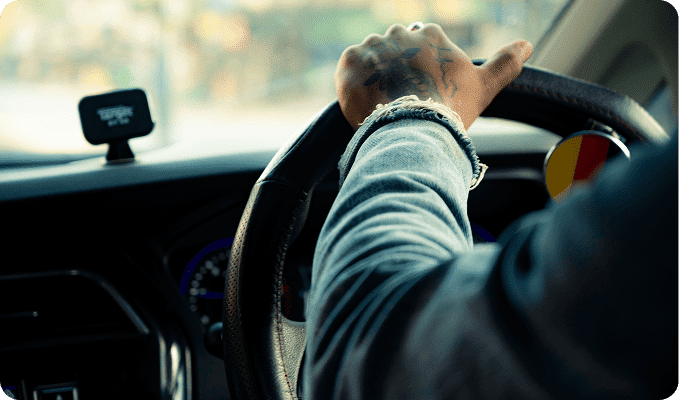 A person with a tattooed hand and denim sleeve grips a car's steering wheel, with a "TECHNOLOGY" screen visible.