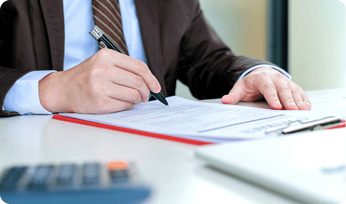 A person in a suit and striped tie signs a document with a black pen on a red clipboard.